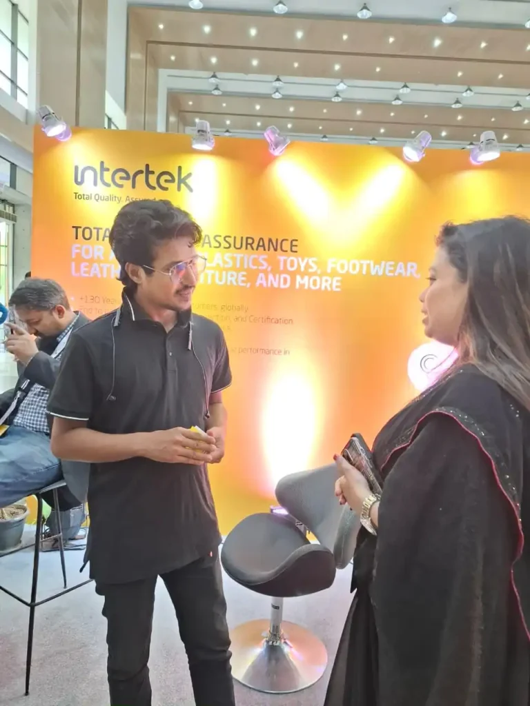 A man discusses quality assurance with a woman at a bright orange Intertek booth featuring product safety messaging.