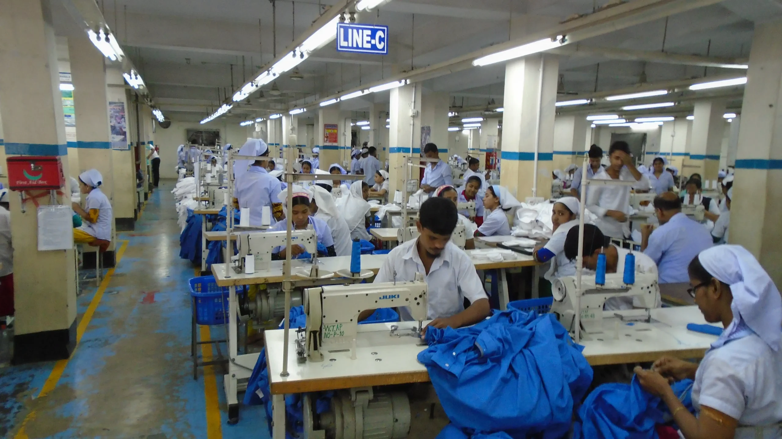 Garment factory sewing floor with skilled operators on industrial machines stitching blue fabric; demonstrates operator skill mapping, inline QC and reduced seam defects. Antor