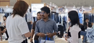 Antor Hossain standing with two visitors at a busy denim sourcing booth, surrounded by hanging jean samples and display tables.