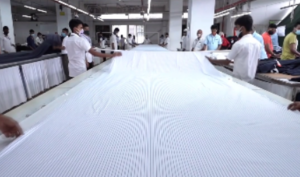 Workers spreading white woven shirting fabric on a long cutting table at a garment factory in Bangladesh